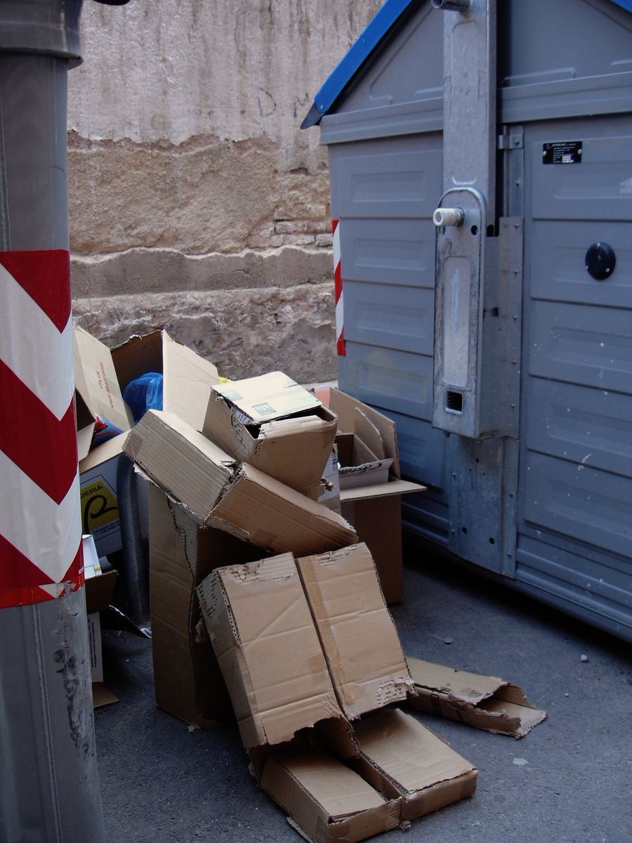 Cardboard boxes arranged into a seated human figure with legs and torso, propped against a stone wall beside grey recycling bins, a red and white striped pole in the foreground.