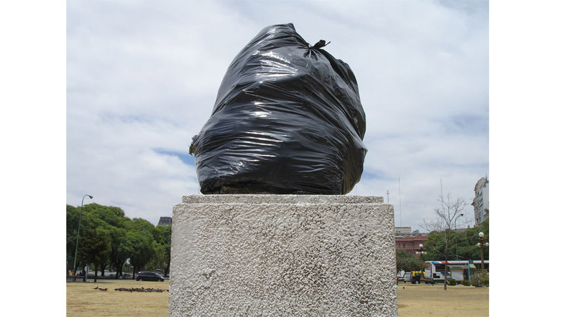 Stone pedestal topped with a large black plastic garbage bag in place of the original bust, park and buildings in the background.
