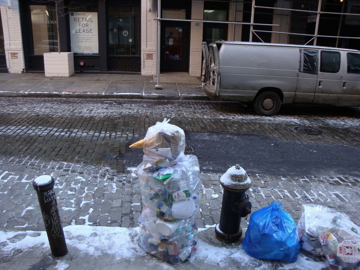 Snowman-like figure made from stacked clear plastic recycling bags full of bottles and cans, beside a fire hydrant on a snowy cobblestone street.