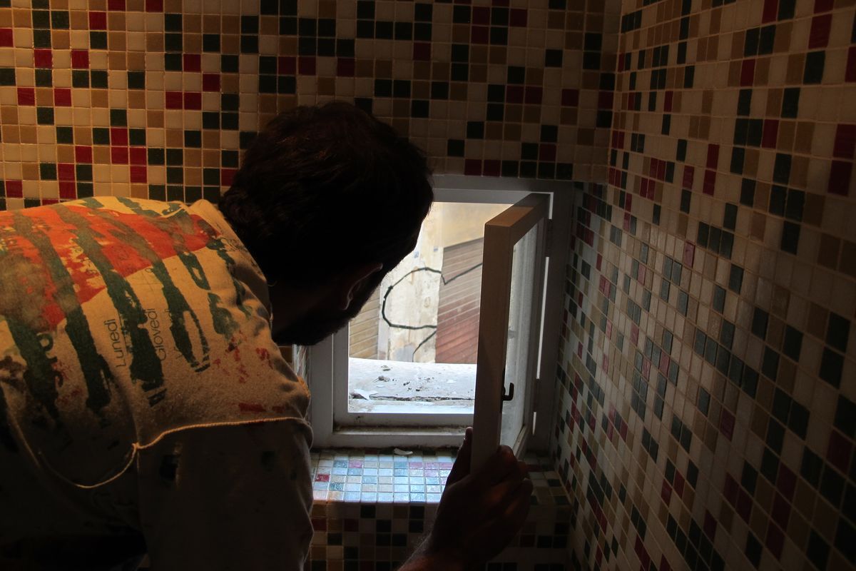 Man leaning through a small tiled window opening in a dim room, looking out at a curved black line painted on a building across a narrow alley.