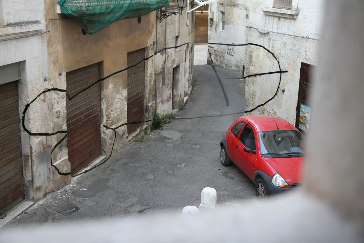 View from above down a narrow alley, a thin curved black line painted across building facades and shutters, a red car parked below.