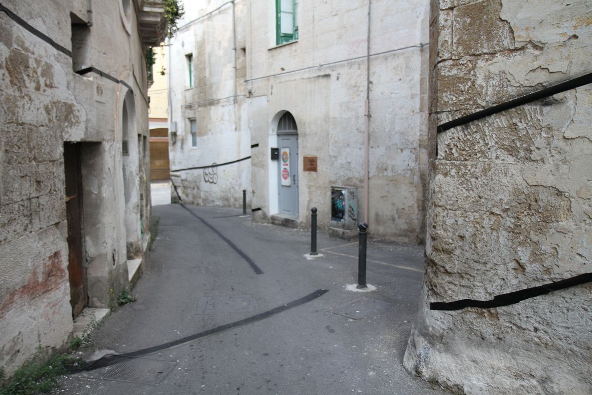 Narrow alley between old stone buildings with a thin black line painted along both walls, leading toward a doorway and bollards.