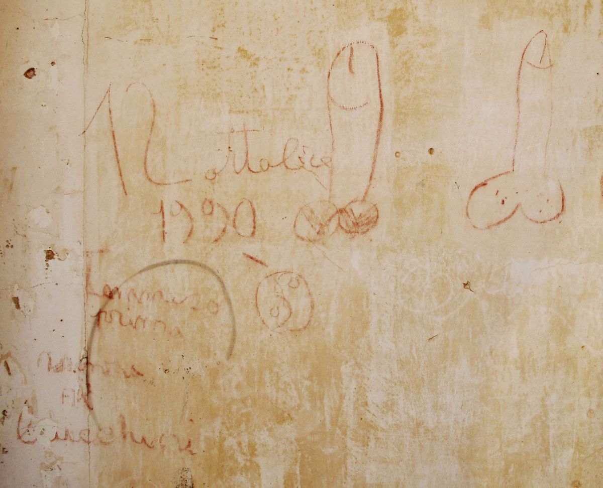 Faded red chalk graffiti on a weathered plaster wall, cursive writing, the date 1990, and crude drawings of genitalia and a circular symbol.