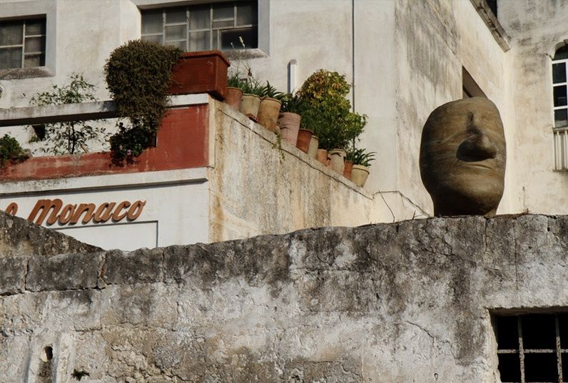 Large egg-shaped clay head sculpture resting on a stone rooftop wall beside a building with potted plants and a sign reading Monaco.