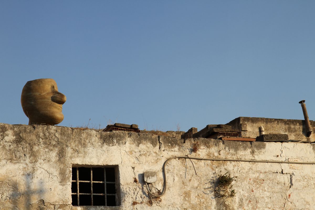 Wide view of an egg-shaped clay head sculpture perched on a weathered rooftop parapet against a clear blue sky, a barred window below.