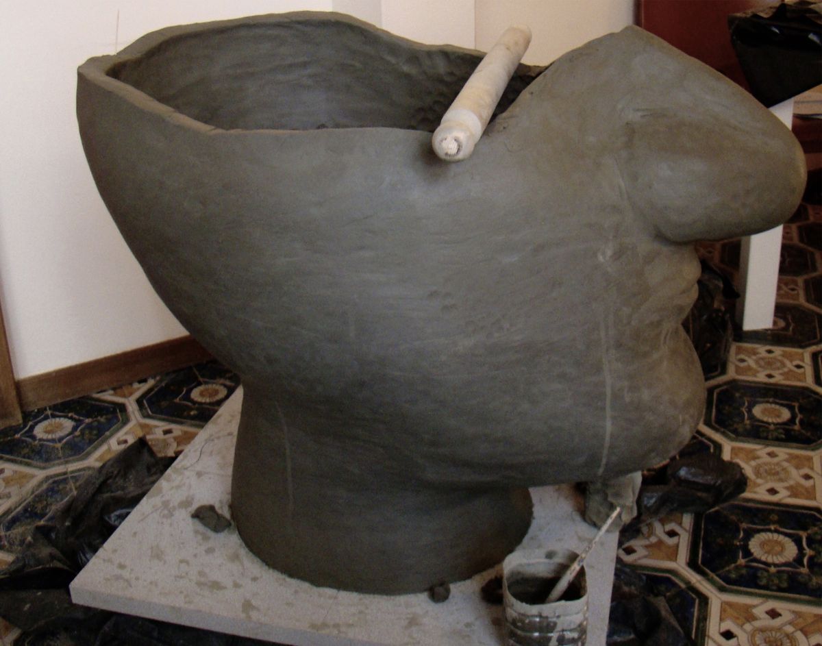 Unfired grey clay bust in progress on an indoor pedestal, a wooden tool resting across the shoulder, tiled floor and buckets nearby.