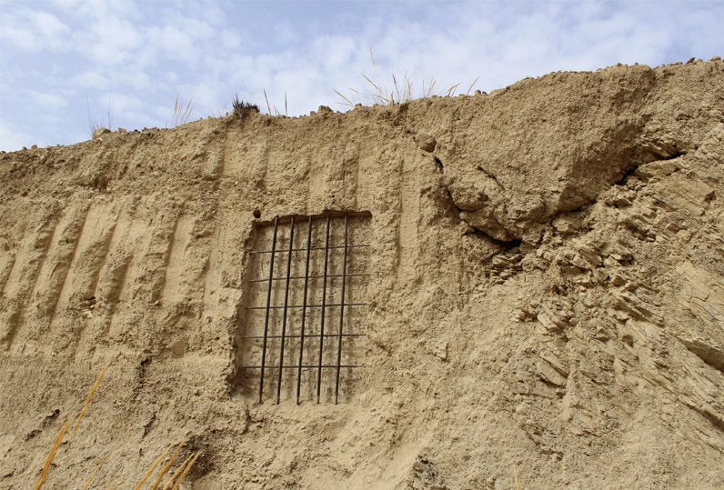 Wide view of a rammed-earth wall with vertical ribbed grooves and a rectangular iron grid window, dry grass at the top edge, cloudy sky above.