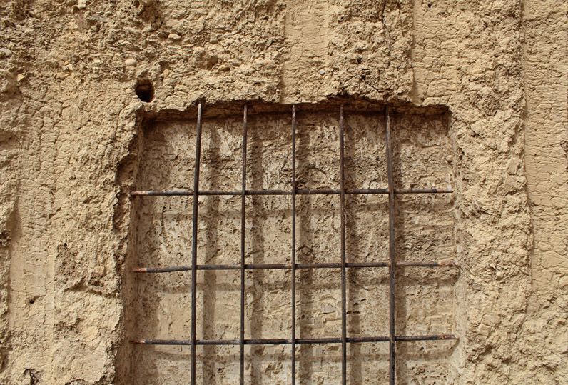 Close-up of a small square iron grid embedded in a rough sand-colored earthen wall, set within a slightly recessed patch of exposed rebar.
