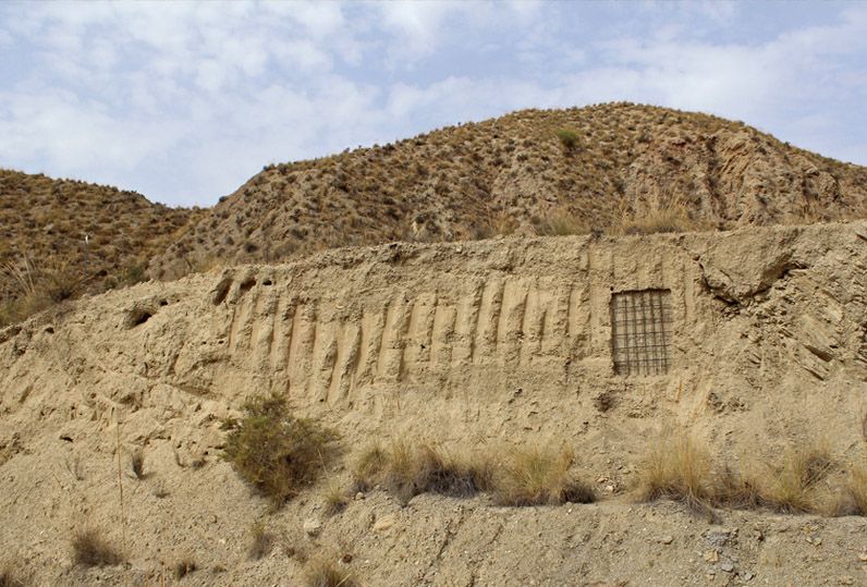 Rammed-earth wall with vertical ribbed grooves and a small embedded iron grid window, arid hills with sparse scrub behind.