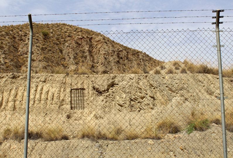 Rammed-earth wall seen through a chain-link and barbed-wire fence, an iron grid window set into the wall, dry hills in the background.
