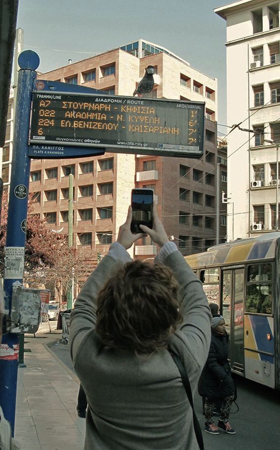 Person photographing an electronic bus timetable sign with a phone, a single bird perched on top, a bus and pedestrians on the street below.