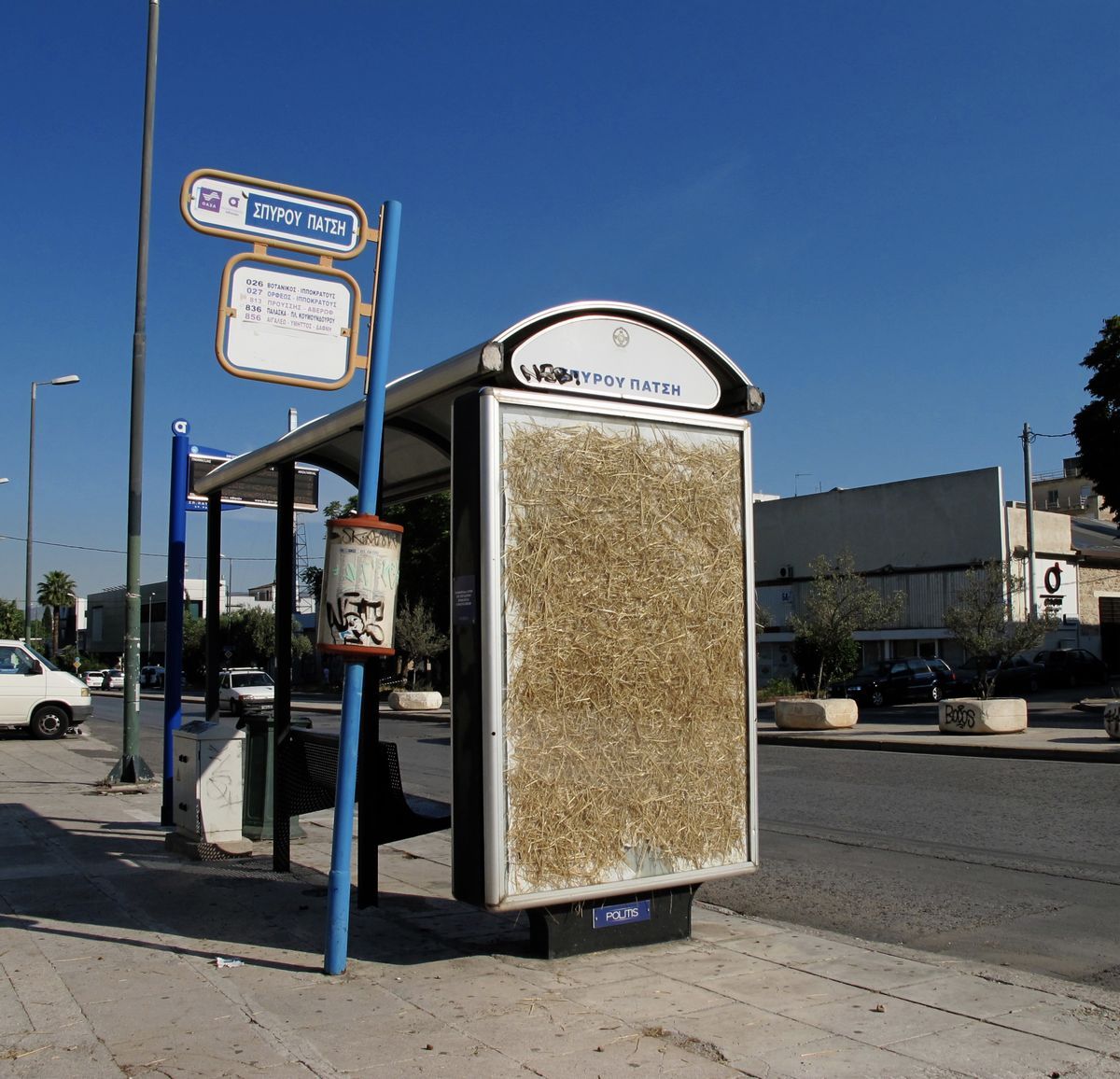 Bus stop shelter with an advertising panel completely filled with loose straw behind its glass, a street sign for the stop beside it under a clear sky.