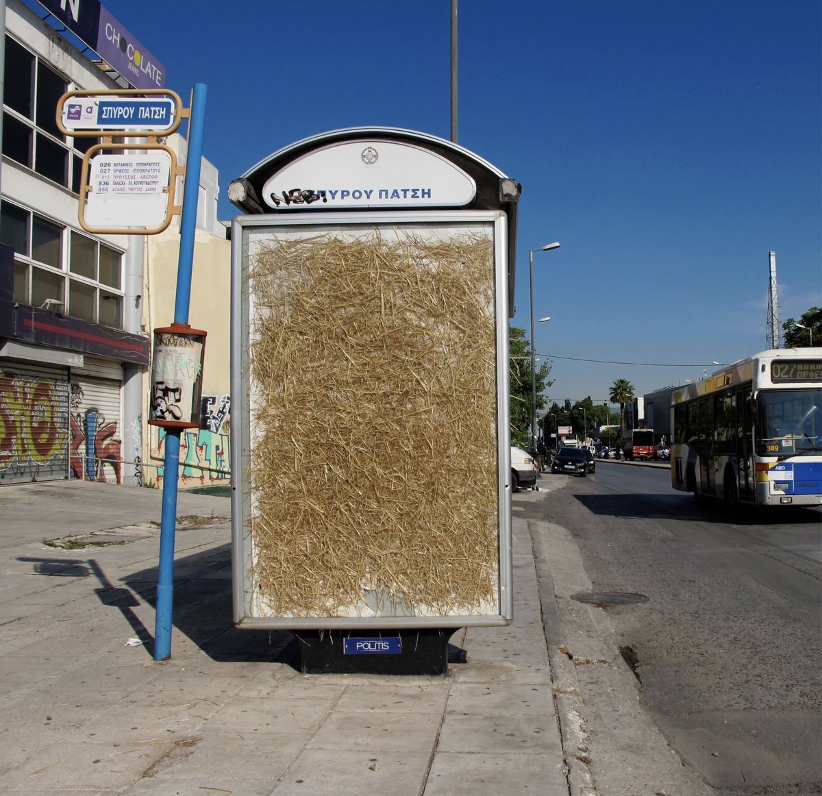 Wide view of a bus stop shelter with its advertising panel filled with straw, graffiti-covered storefronts and a city bus passing on the street.