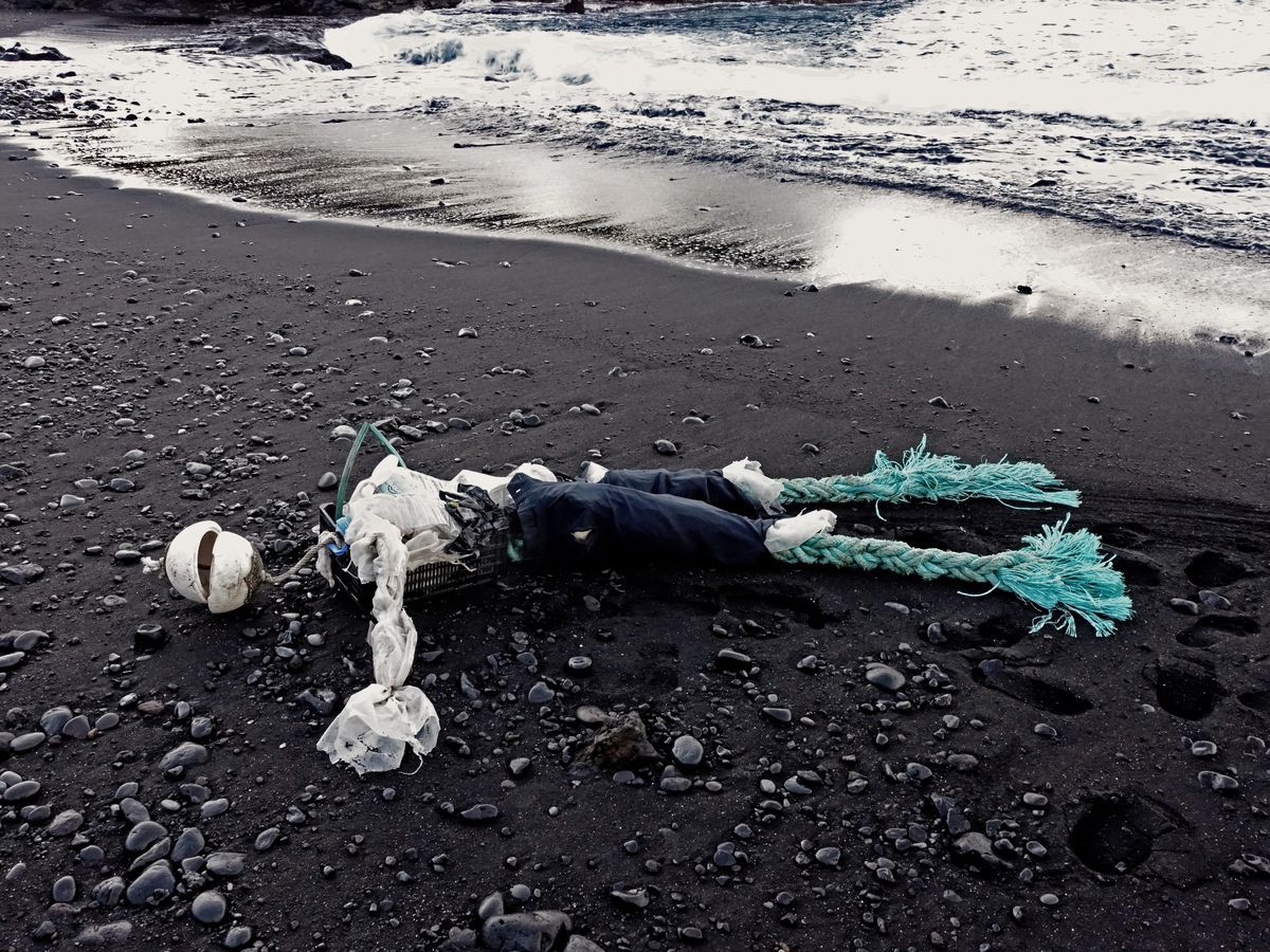 Human figure lying on black volcanic sand assembled from beach debris, a white buoy for a head, plastic bags and rubbish for a torso, two braided teal rope legs, waves breaking behind.