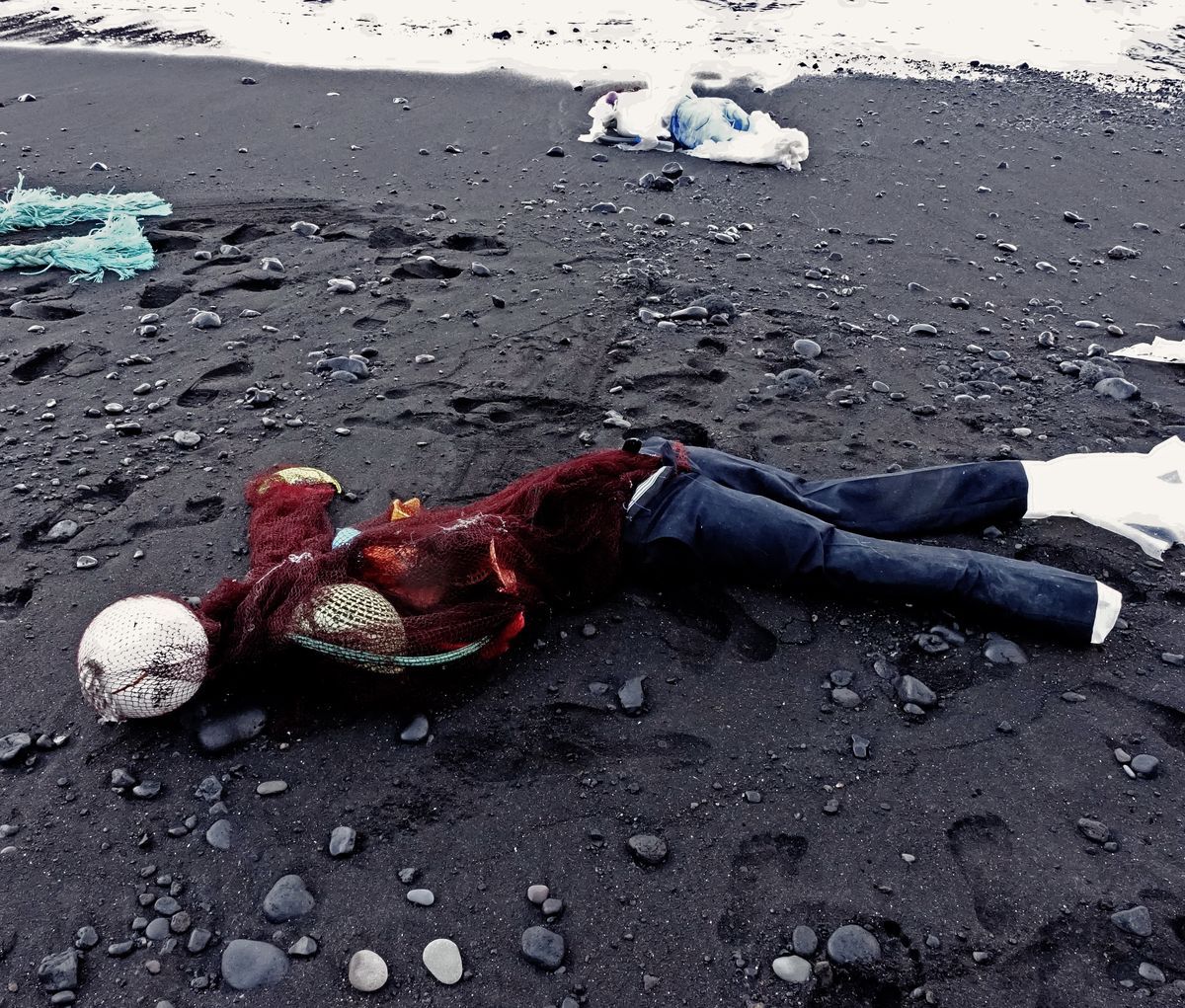 Human figure lying on black sand made from a red fishing net torso with mesh buoy accents, denim-colored plastic legs, a white plastic bundle nearby, waves in the background.