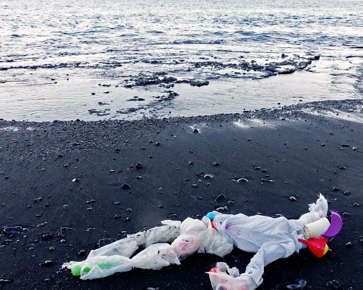Human figure lying on black sand near the waterline formed from white plastic sheeting and bags, its head made of a stacked colorful cup, lid, and funnel, calm water behind.