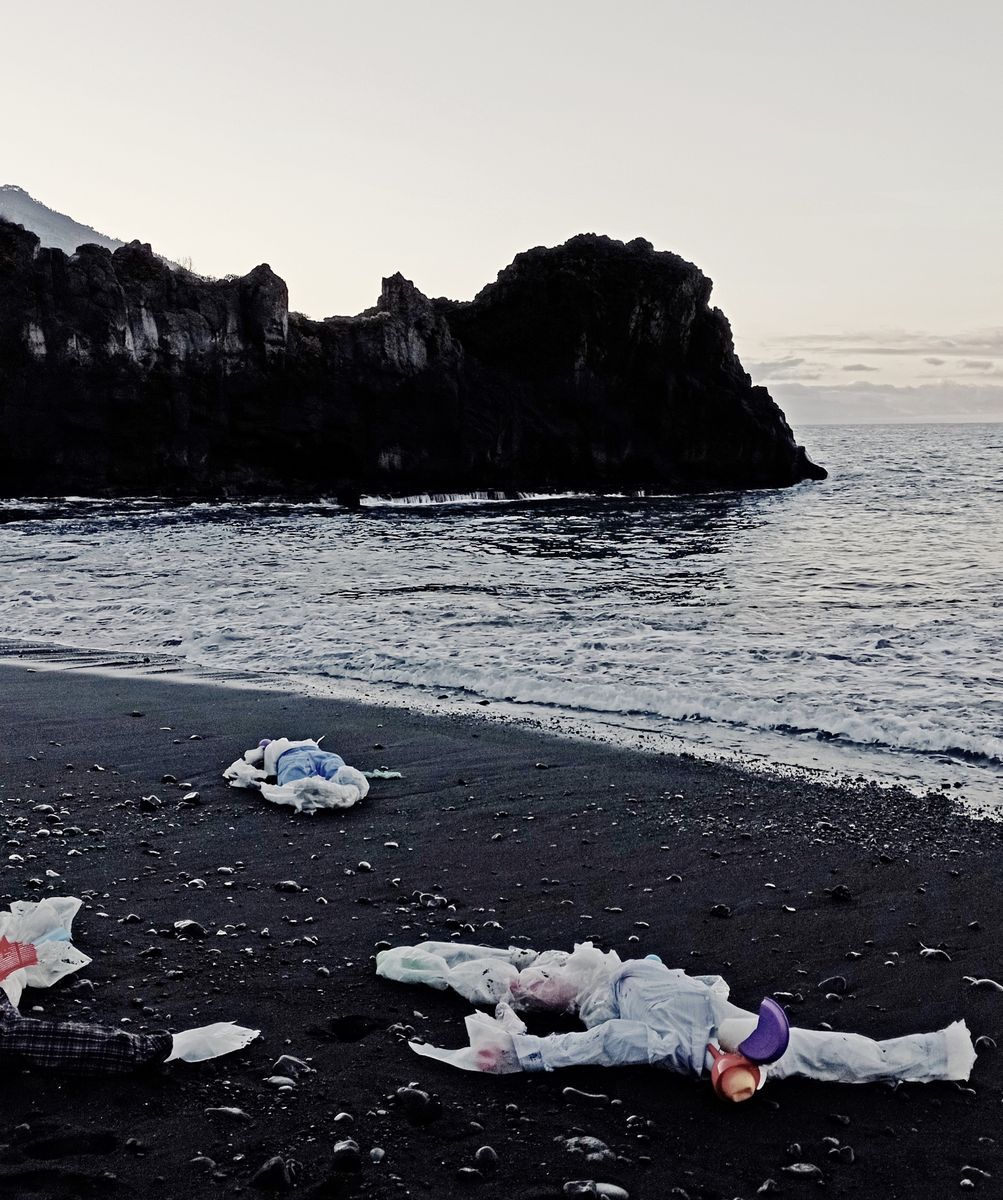Wide view of a black sand beach with two reclining human figures made of white plastic debris scattered near the shoreline, dark sea cliffs and calm water in the background.