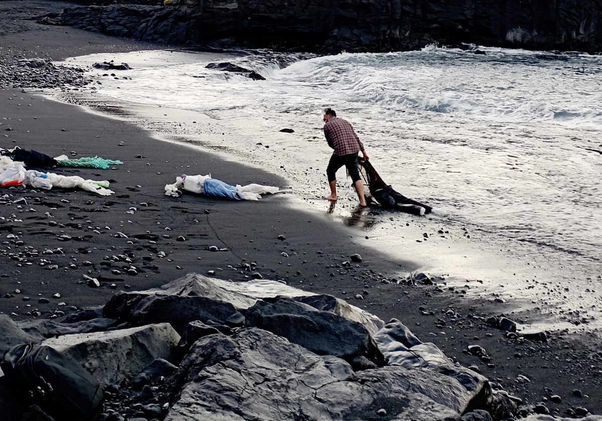 Man in a plaid shirt crouching at the water's edge on a black sand beach, pulling a dark plastic figure from the surf, scattered debris and rope nearby.