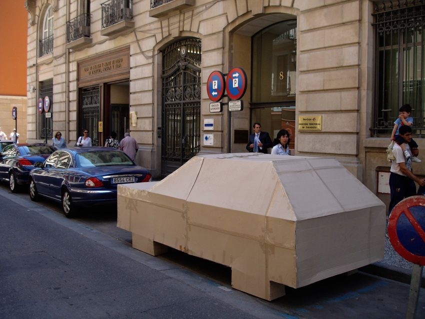 Full-size cardboard car sculpture parked at the curb outside a stone building, real cars and pedestrians on the street beside it.