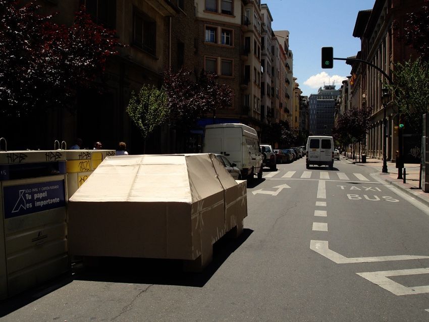 Cardboard car sculpture parked beside recycling bins on a city street, vans and buildings receding down the road.
