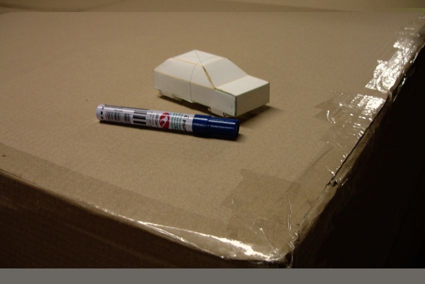 Small white paper model of a car beside a blue marker pen, resting on a cardboard box corner.