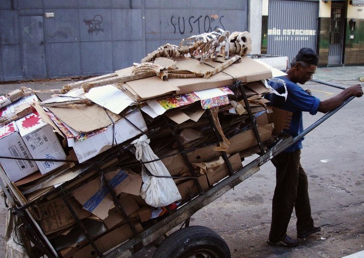 Man pulling a heavily loaded cardboard recycling cart through a street, a large cardboard skeleton sculpture strapped on top.