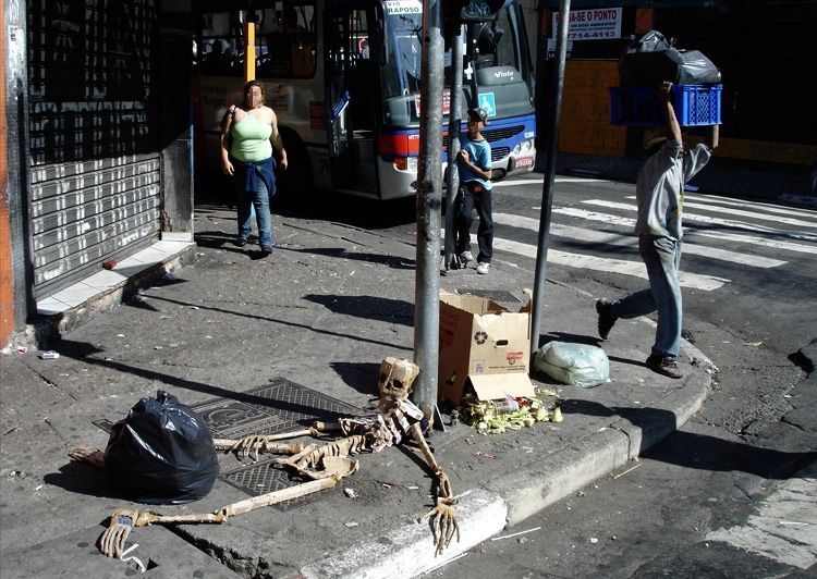 Cardboard skeleton sculpture lying on a street corner beside boxes and trash bags, pedestrians and a bus passing behind.
