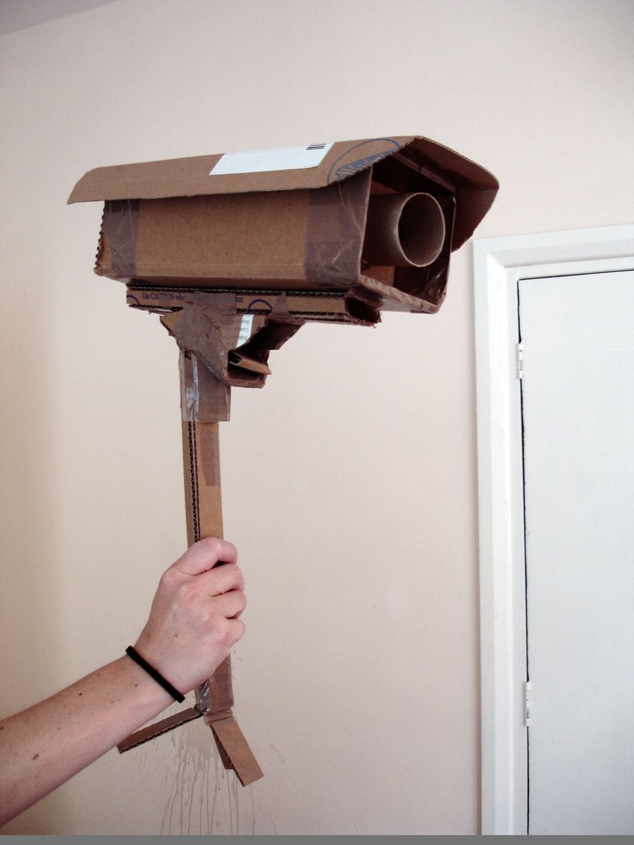 Hand holding up a handmade cardboard surveillance camera model against a plain wall, next to a white door.