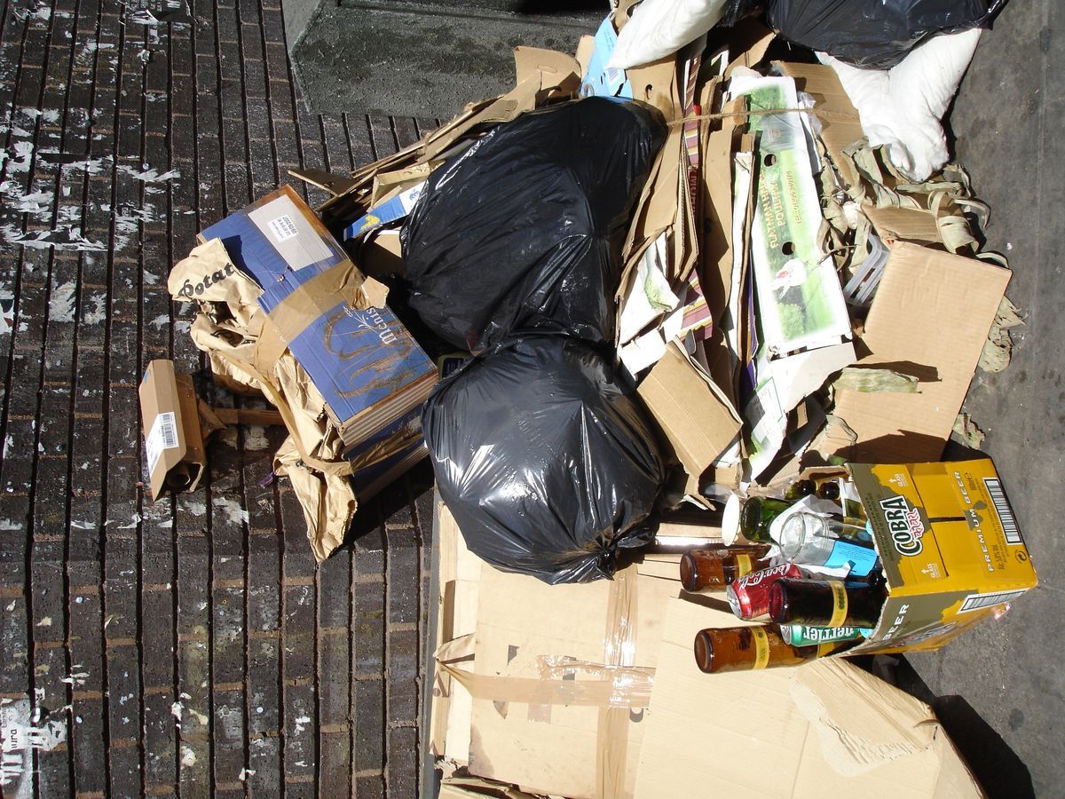 Cardboard surveillance camera camouflaged among a pile of rubbish, black bin bags, flattened boxes, and beer bottles on a brick pavement.