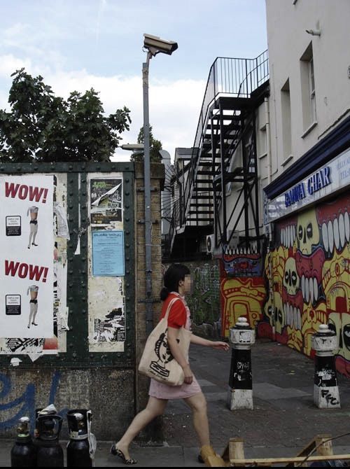 Cardboard surveillance camera mounted on a tall pole above a graffiti-covered wall, a woman walking past on the street below.