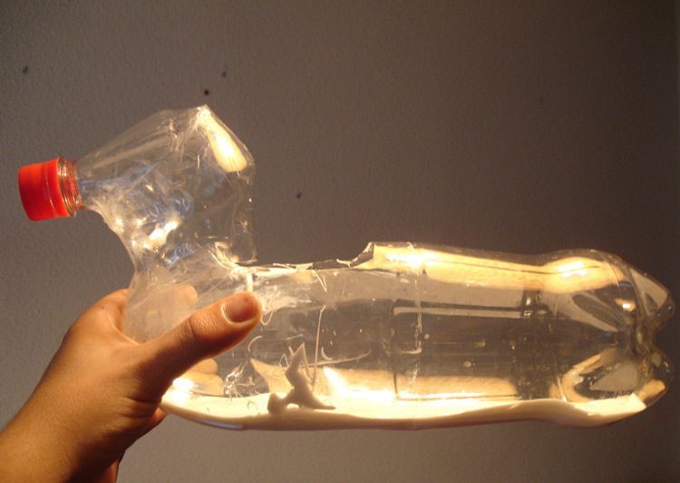 Close-up of a hand holding a swan-shaped decoy made from a clear plastic bottle with a red cap, backlit against a plain wall.
