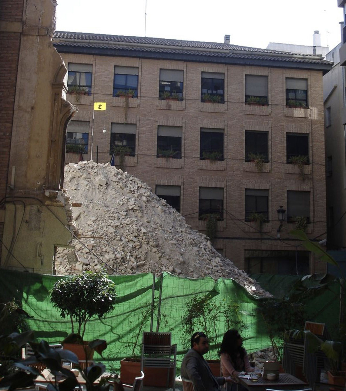 A large mound of pale rubble and debris piled against a brick apartment building, seen from a green-fenced outdoor café terrace with diners at tables in the foreground.