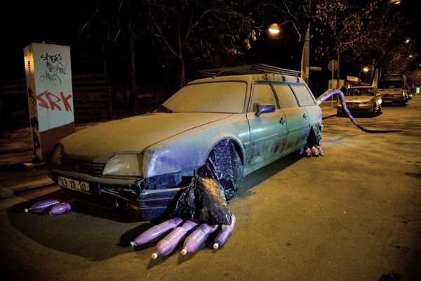 Abandoned station wagon at night disguised as a rat, a long tail made from a purple tube trailing from the rear, clawed paws made from bundles of bottles wrapped in dark plastic bags beneath the front wheel.