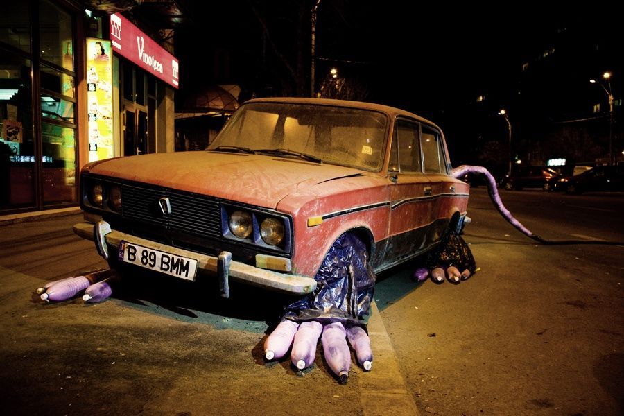 Old red sedan disguised as a rat, clawed paws made from bundles of bottles in dark plastic bags emerging from under both front and rear wheels, a long purple tail trailing off to the right.