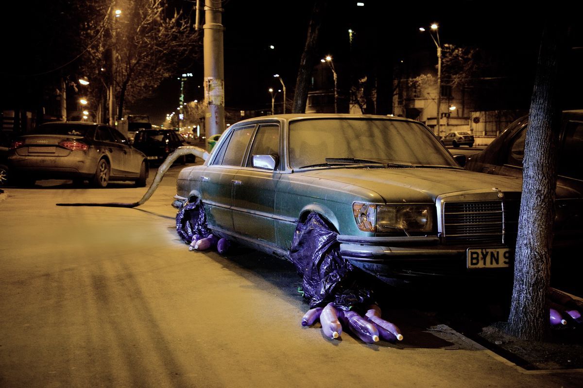 Vintage Mercedes sedan disguised as a rat, clawed paws made from bundles of bottles in dark plastic bags spilling from under the front wheel, a long purple tail stretching away down the sidewalk.