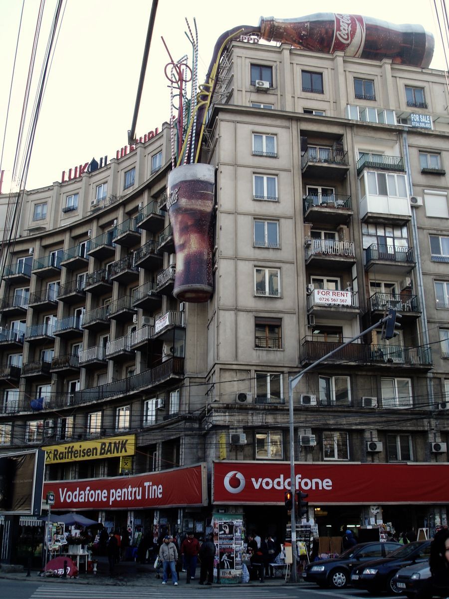 Curved building facade with an oversized Coca-Cola bottle and glass mounted on the rooftop and side wall, bank and phone company signage below.