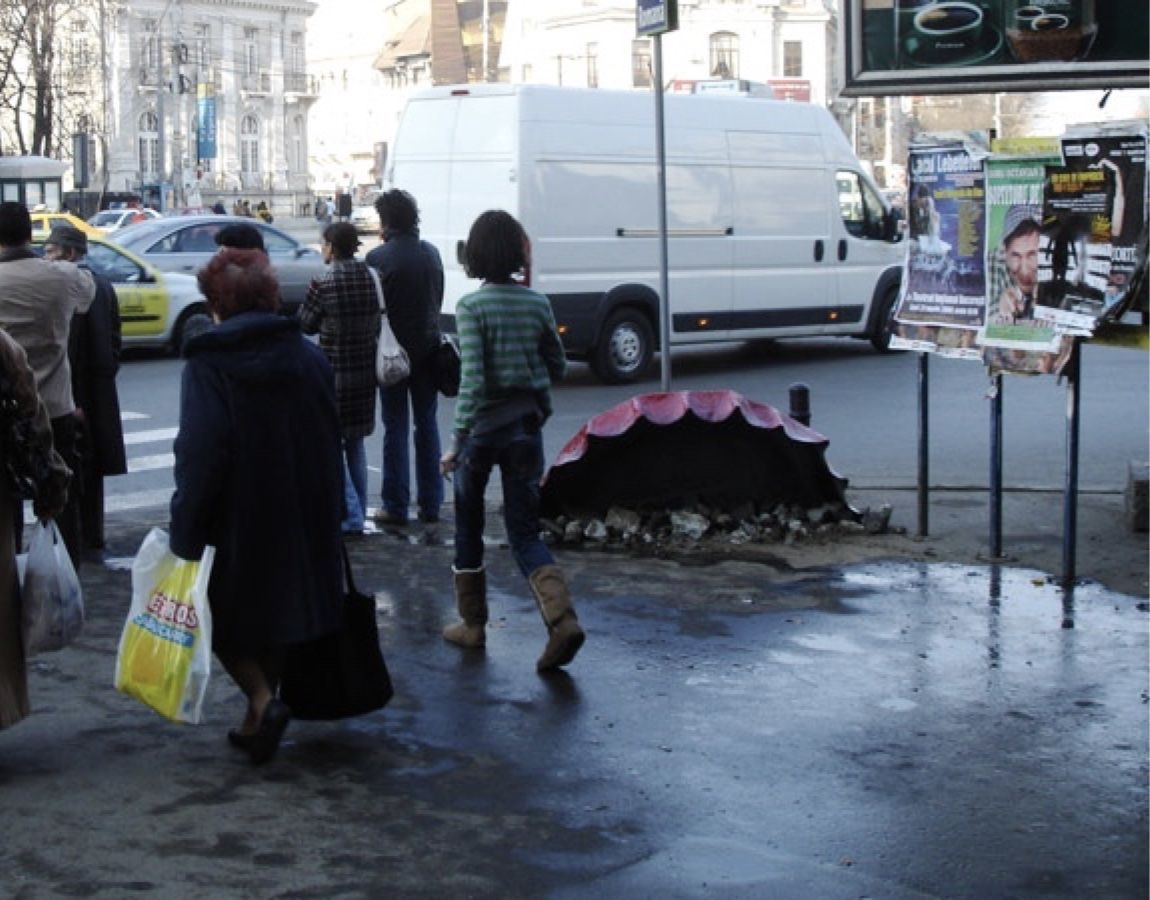Street scene with pedestrians and a van passing a large pink and black shell-shaped object resting on the pavement beside a curb.