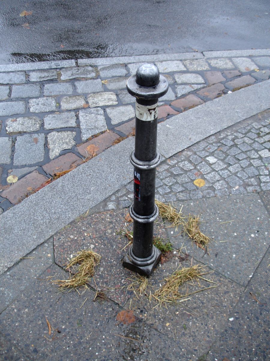 Black bollard on a cobblestone street corner with loose straw scattered around its base, wet pavement and stone curb nearby.