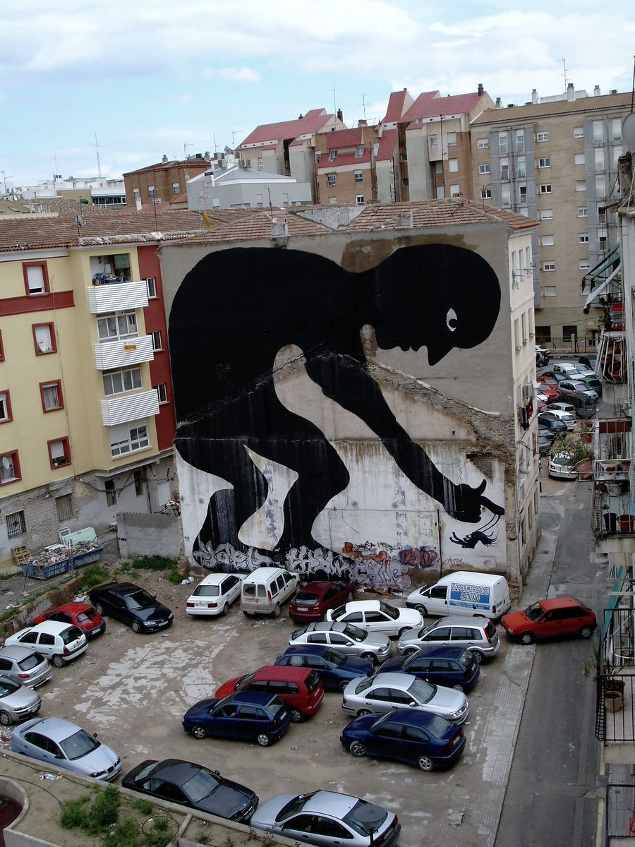 Giant black silhouette of a crouching figure bent over a rooftop edge, one hand reaching down to hold a leash attached to a small dog, apartment buildings and a parking lot with rows of cars below.