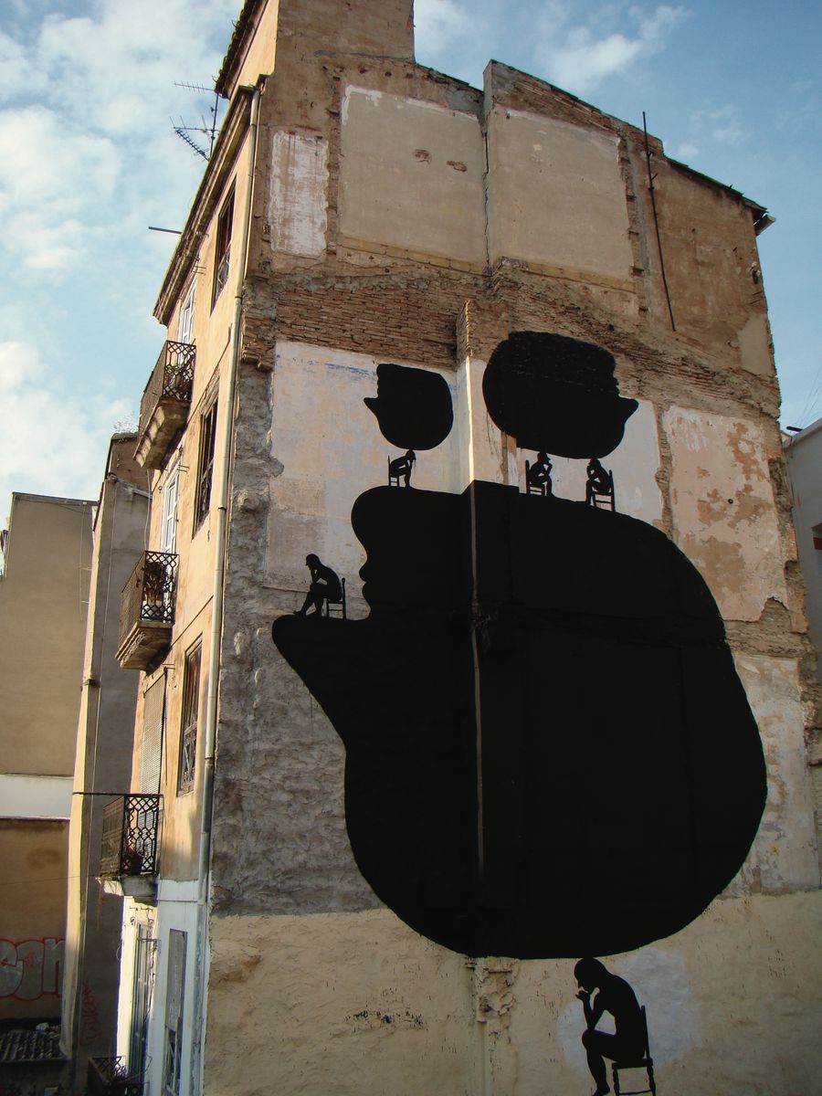Tall building corner painted with a large black silhouette of a hand and face profile, with three small seated figures on chairs perched along the fingertips, thinking poses, balconies on the building beside it.