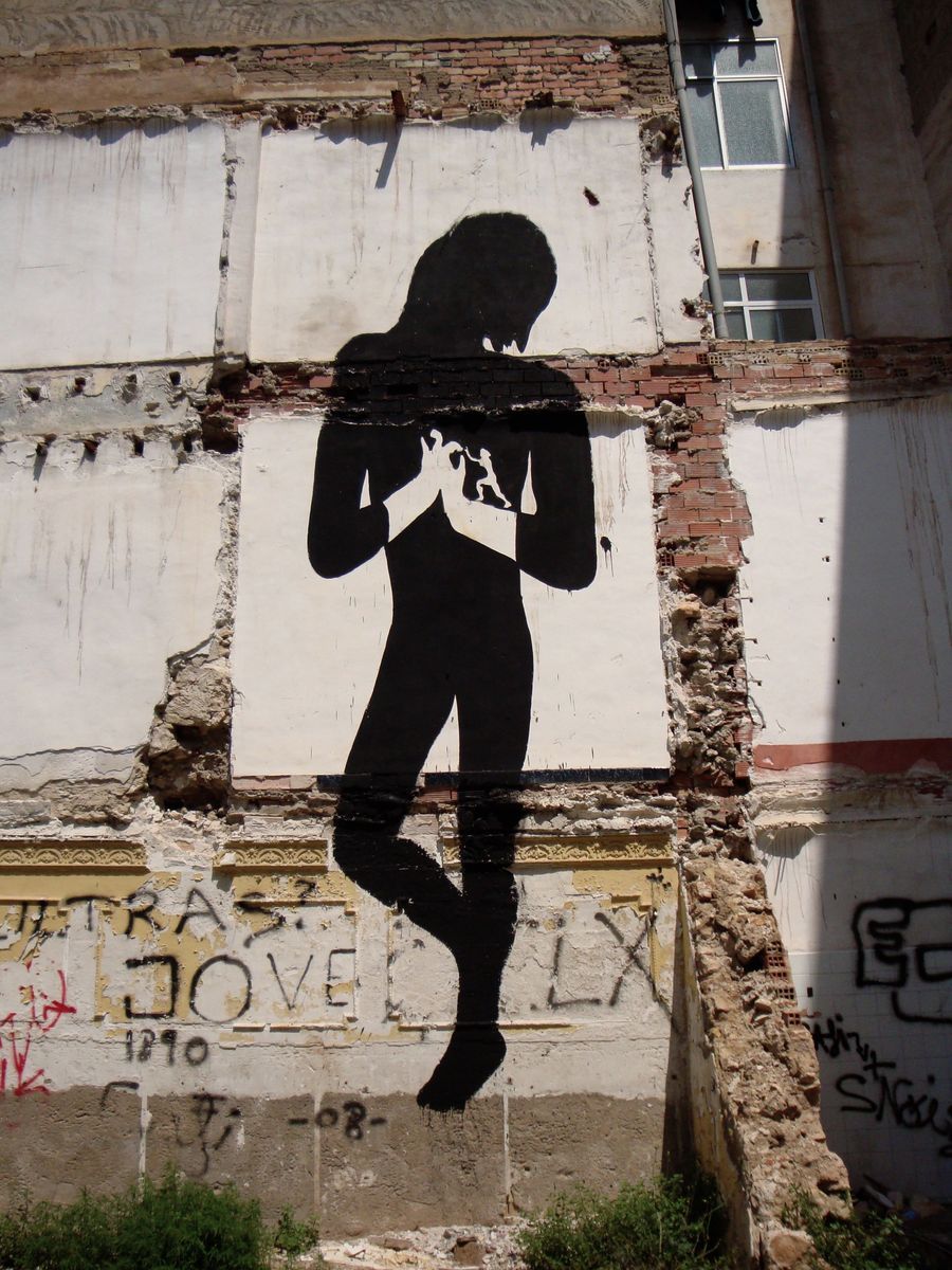 Black silhouette figure holding two hands together in front of its chest, cradling a small white heart shape, painted on a square white panel set into a weathered brick and plaster wall.