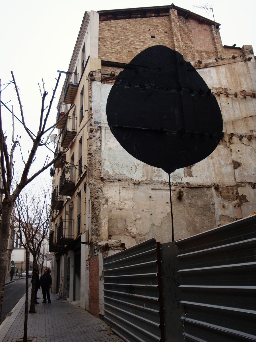Large black egg-shaped silhouette suspended by a thin black line above a corrugated metal fence, painted on the corner of a weathered stone building, bare trees and pedestrians on the street below.