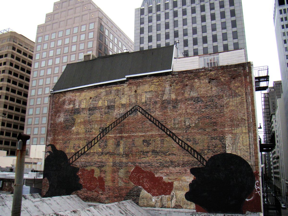 Old brick warehouse wall with two black silhouette profile heads facing each other at roof level, connected by a black zigzag ladder shape, skyscrapers surrounding the building.