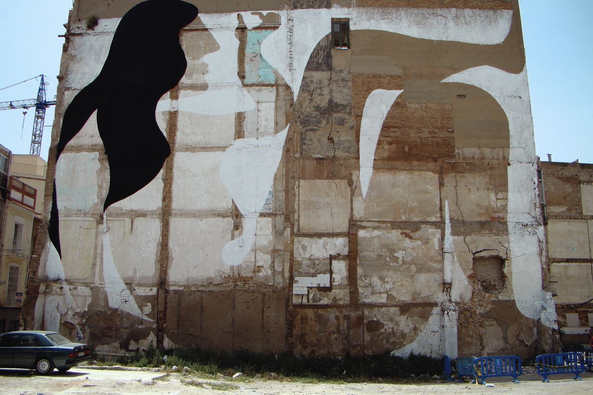 Large abstract black and white shapes painted across a weathered stone building facade under demolition, a construction crane visible behind, a parked car and blue barriers in front.
