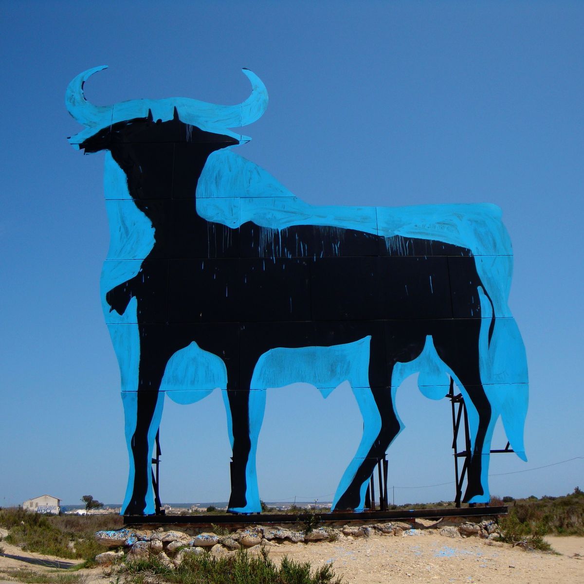 Giant black silhouette of a bull outlined in light blue standing in an open field under a clear sky, mounted on a metal support structure.