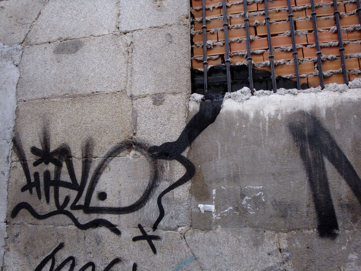 Small black fish graffiti tag spray-painted on a stone wall beside a bricked-up barred window, a black drip trailing down from the sill.