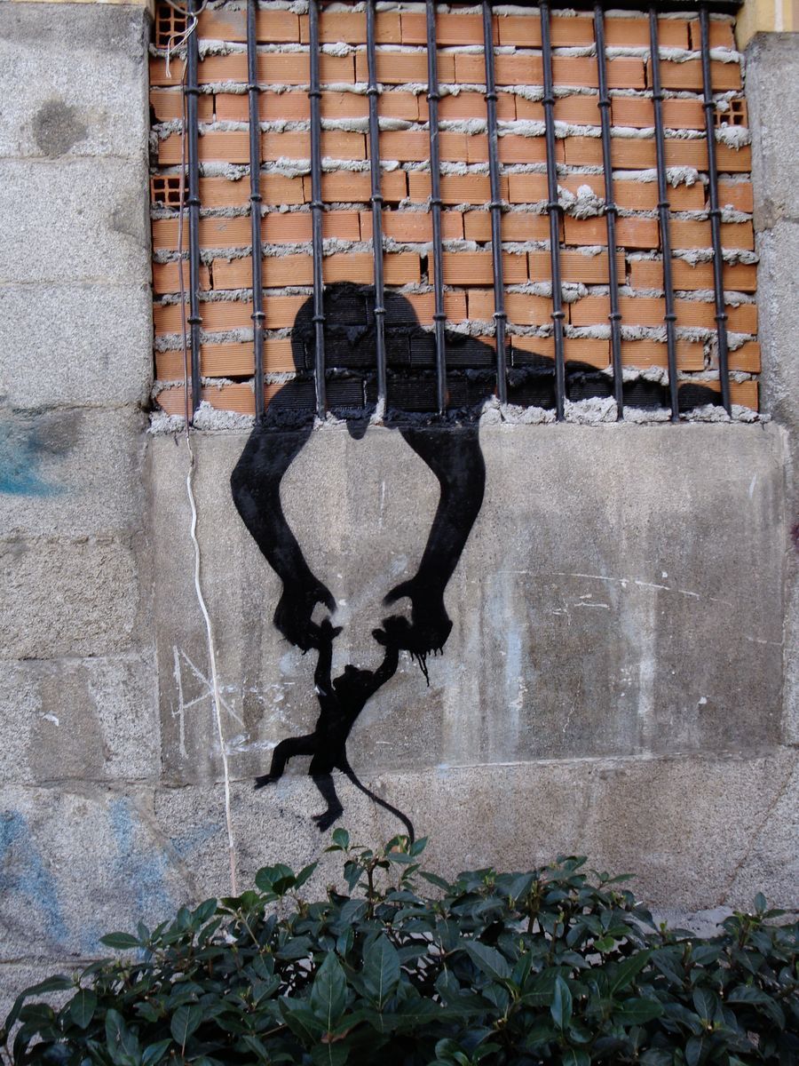 Black silhouette figure with arms reaching down through a barred window, holding a small monkey by its paws, green bushes at the base of the wall.