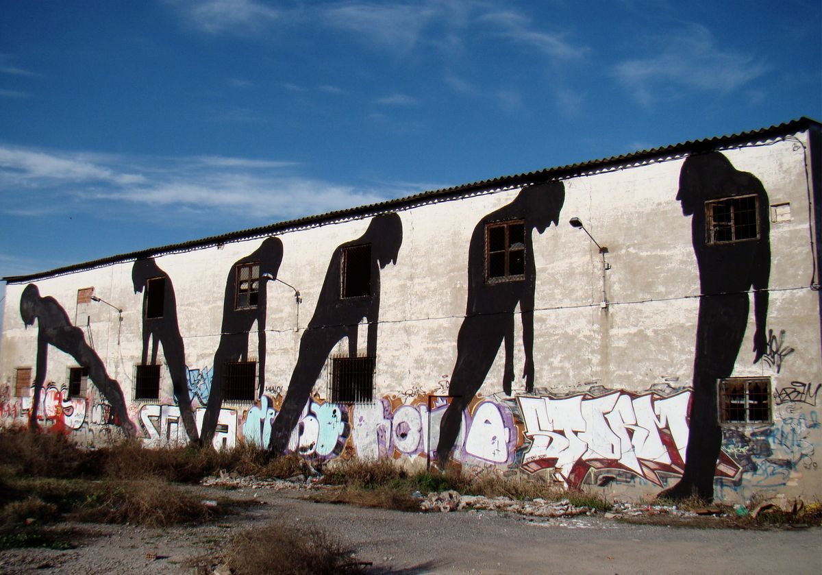 Row of hunched black silhouette figures shuffling forward across a long warehouse facade, barred windows and a spray-painted graffiti tag along the base of the wall.