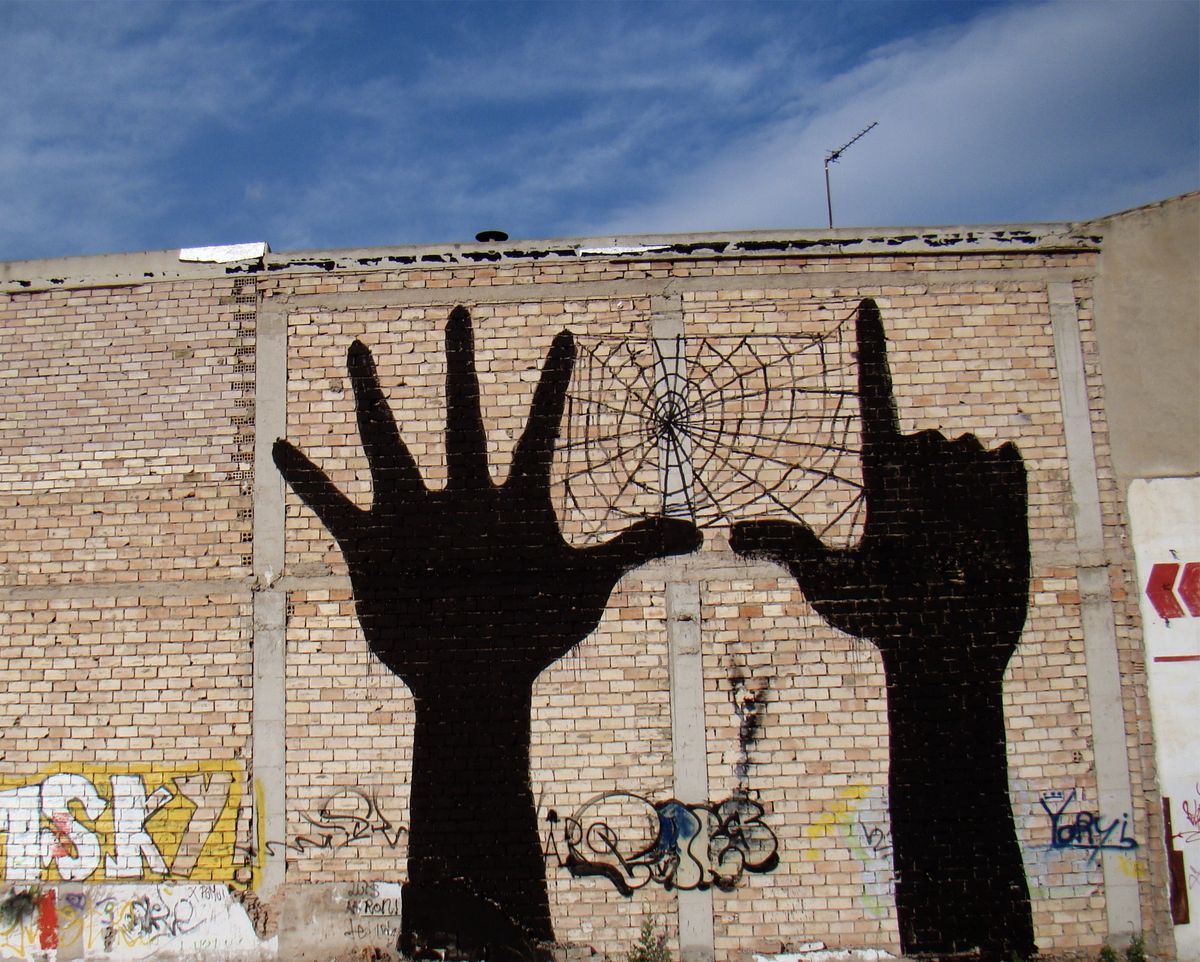 Two large black silhouette hands reaching up from a brick wall, a spider web painted between their fingertips, graffiti tags scattered around the base.