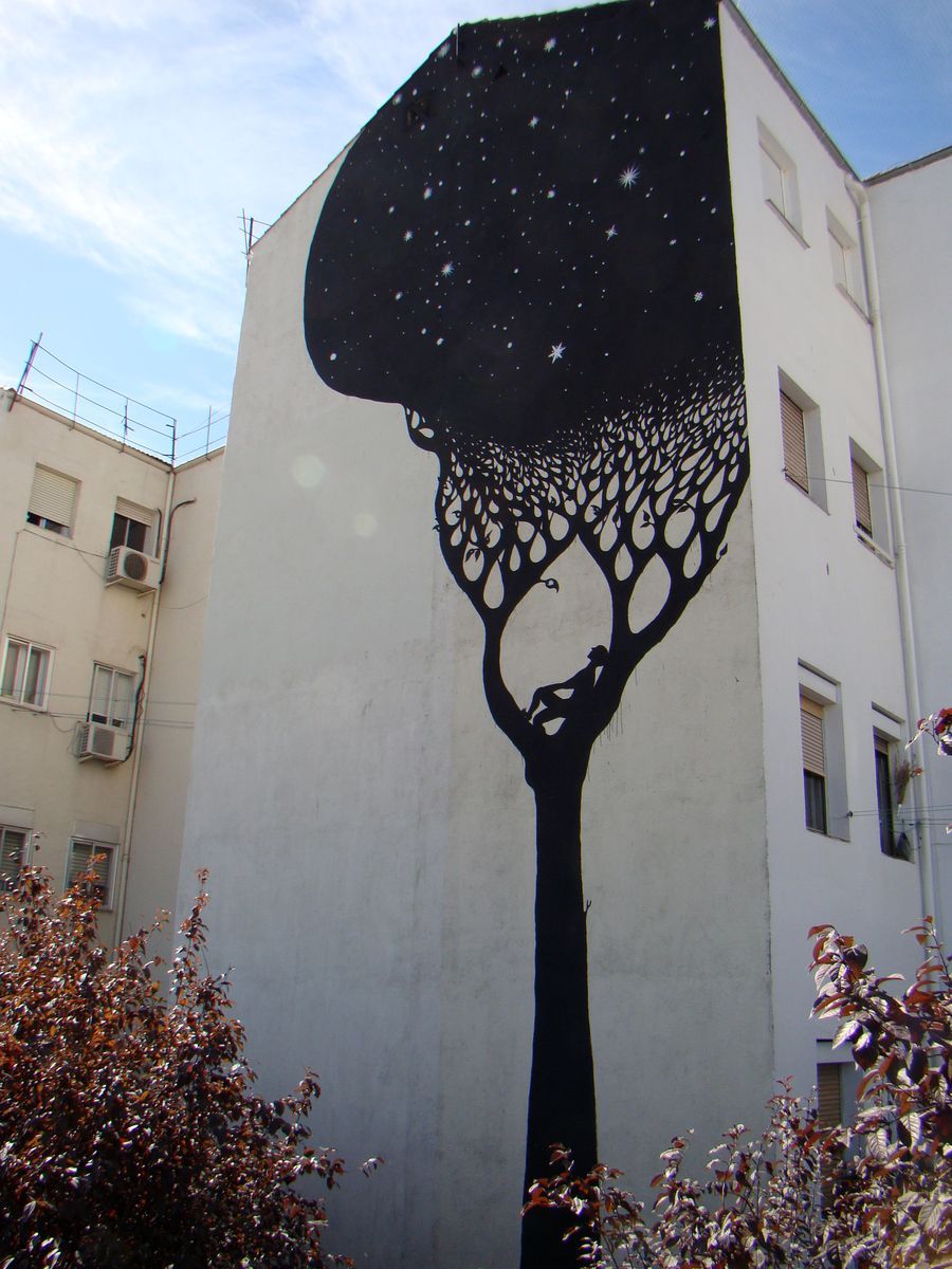 Full building gable painted with a tall black tree silhouette against a starry sky, its canopy made of teardrop-shaped leaves, a small figure resting in a hollow near the base, apartment windows on either side.